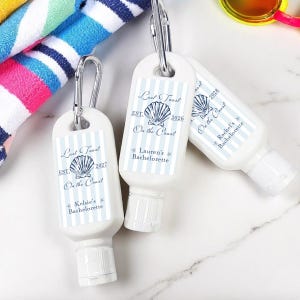 May include: White sunscreen bottles with silver carabiner clips. Each bottle has a blue and white striped label with text that reads "Last Toast On the Coast" and a seashell graphic. The bottles are labeled with names and dates.