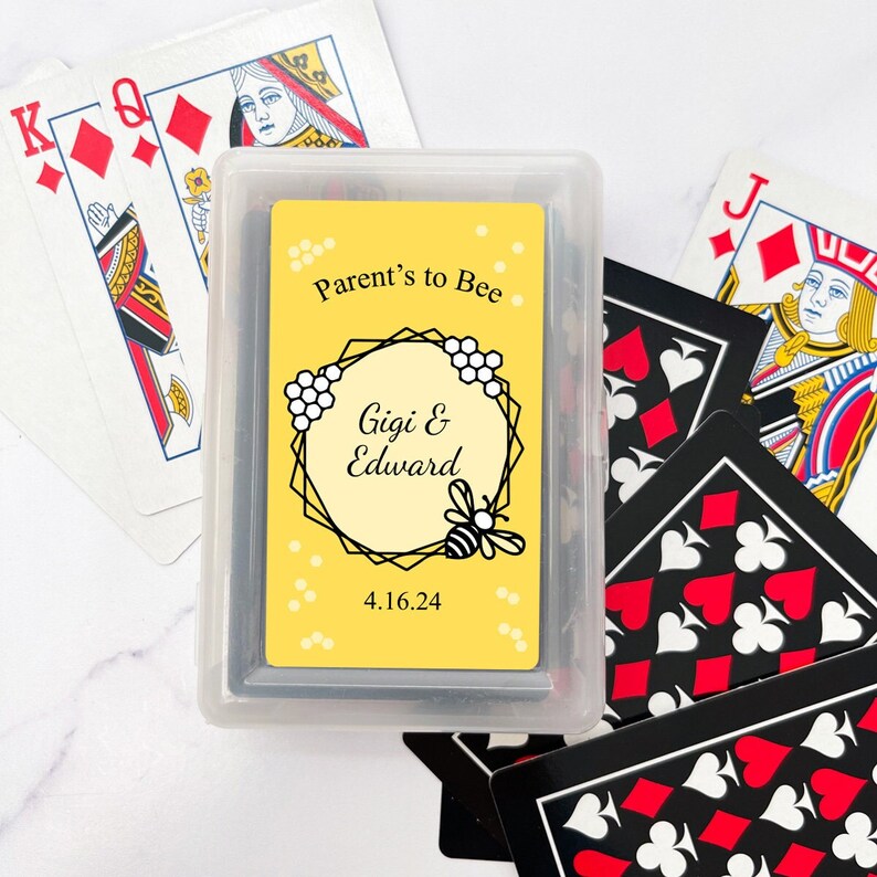 May include: A deck of playing cards in a clear plastic case. The card design features a yellow background with the text "Parent's to Bee," the names "Gigi & Edward," and the date 4.16.24, along with a bee and honeycomb design. Other playing cards are scattered around.