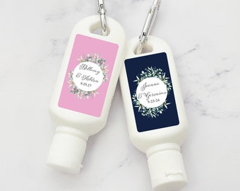 May include: Two white hand sanitizer bottles with silver carabiner clips. One has a pink label with a floral wreath and the names "Bellamy & Ashton" and the date "9.25.27." The other has a navy label with "Joanna & Geronimo" and "5-23-24."