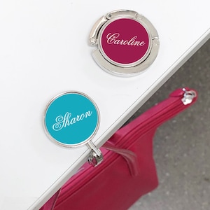 May include: Two silver-toned handbag hooks with personalized names. The top hook has a fuchsia-colored disc with the name "Caroline" in white script. The bottom hook has a turquoise-colored disc with the name "Sharon" in white script.