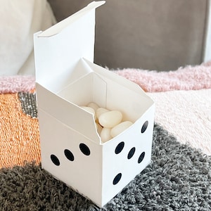 May include: A white cardboard box shaped like a dice with black dots. The box is open and filled with white jelly beans.