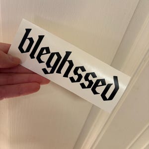 May include: A white sticker with the word "bleghssed" printed in black gothic font. The sticker is rectangular and held by a hand against a white background.