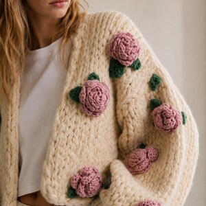 May include: Cream-colored, chunky knit cardigan adorned with multiple pink rose appliqu&eacute;s and green leaves. The sweater has a relaxed fit and long sleeves. A white t-shirt is visible underneath.