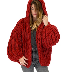 Op de afbeelding: Een grof gebreid vest in een levendige rode kleur met een oversized capuchon, lange volumineuze mouwen en een dik, getextureerd kabelbreipatroon. Het kledingstuk wordt gedragen over een donkergrijs t-shirt en een donkergrijze spijkerbroek.