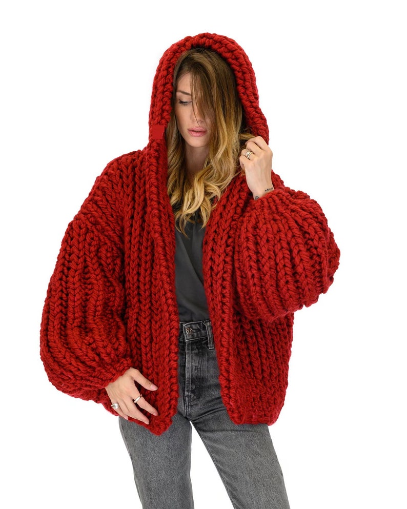 Op de afbeelding: Een grof gebreid vest in een levendige rode kleur met een oversized capuchon, lange volumineuze mouwen en een dik, getextureerd kabelbreipatroon. Het kledingstuk wordt gedragen over een donkergrijs t-shirt en een donkergrijze spijkerbroek.