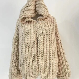 Op de afbeelding: Een beige vest met capuchon gemaakt van dik, grof gebreid garen. Het kledingstuk heeft een opvallende verticale ribbelstructuur over het hele lichaam en de mouwen, met lange, losvallende mouwen en een grote, gestructureerde capuchon bij de hals.