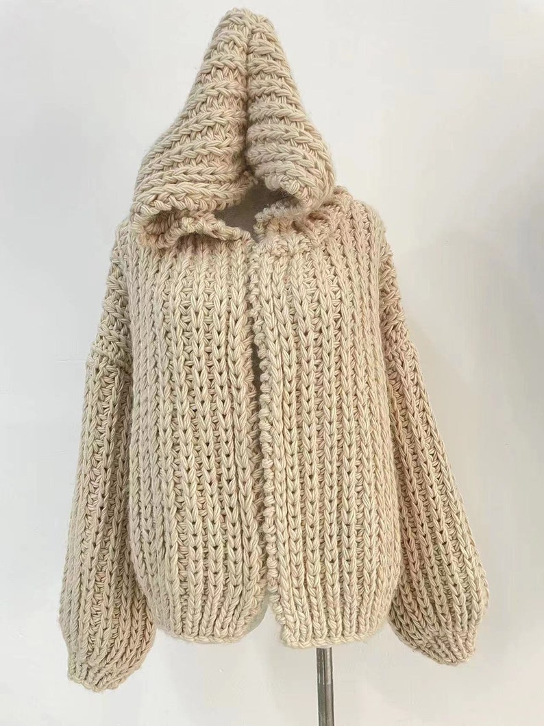 Op de afbeelding: Een beige vest met capuchon gemaakt van dik, grof gebreid garen. Het kledingstuk heeft een opvallende verticale ribbelstructuur over het hele lichaam en de mouwen, met lange, losvallende mouwen en een grote, gestructureerde capuchon bij de hals.