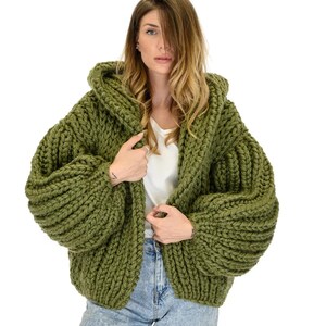 Op de afbeelding: Een olijfgroen grofgebreid vest met een capuchon, lange volumineuze mouwen met geribbelde boorden en een dikke, getextureerde garenconstructie. Het kledingstuk wordt gedragen over een effen wit topje en een lichtblauwe spijkerbroek met grof breipatroon.
