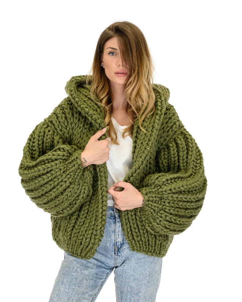 Op de afbeelding: Een olijfgroen grofgebreid vest met een capuchon, lange volumineuze mouwen met geribbelde boorden en een dikke, getextureerde garenconstructie. Het kledingstuk wordt gedragen over een effen wit topje en een lichtblauwe spijkerbroek met grof breipatroon.