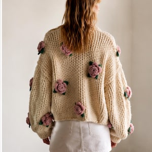 May include: Cream-colored, chunky knit sweater adorned with multiple pink and green floral appliqu&eacute;s. The sweater has a high neckline and long sleeves. The model is wearing a white denim skirt.