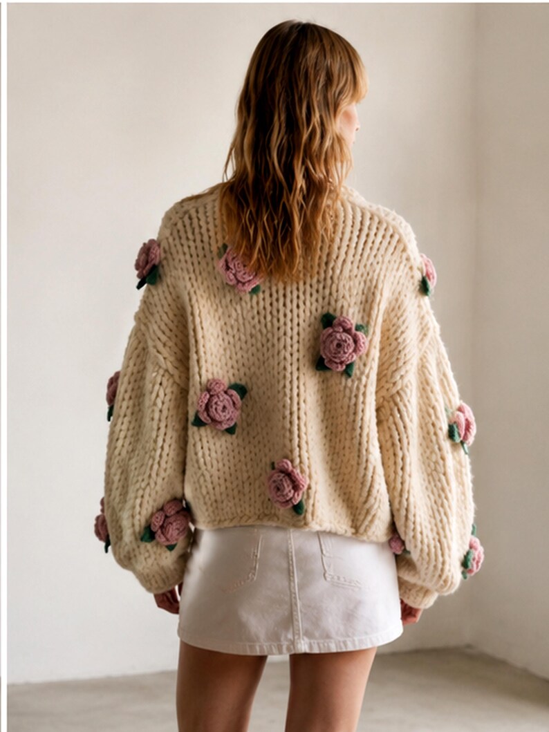 May include: Cream-colored, chunky knit sweater adorned with multiple pink and green floral appliqu&eacute;s. The sweater has a high neckline and long sleeves. The model is wearing a white denim skirt.