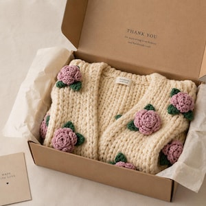 May include: Cream-colored, chunky knit cardigan adorned with crocheted pink roses and green leaves, presented in a brown cardboard box. The box includes a small card that says "Made with Love". The cardigan is a handmade item.