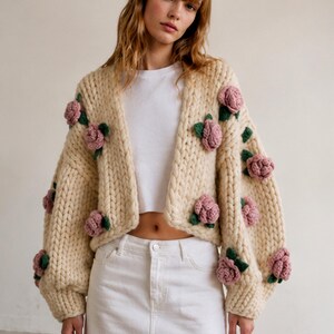 May include: Cream-colored, chunky knit cardigan adorned with pink rose appliqu&eacute;s and green leaves. The open-front sweater has long sleeves and is a statement piece. The cardigan is a unique, handmade garment.