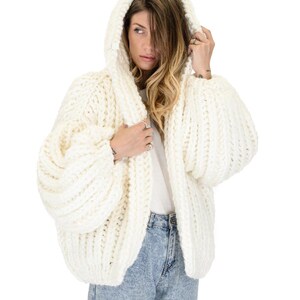 Op de afbeelding: Een gebroken wit, grof gebreid vest met capuchon, voorzien van een dikke ribbelstructuur en oversized, volumineuze mouwen. Het kledingstuk wordt gedragen over een effen wit t-shirt en een lichtblauwe, acid-wash spijkerbroek met grove textuur.