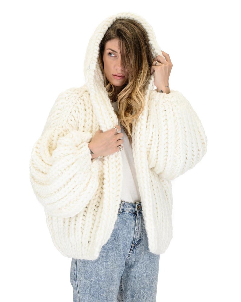 Op de afbeelding: Een gebroken wit, grof gebreid vest met capuchon, voorzien van een dikke ribbelstructuur en oversized, volumineuze mouwen. Het kledingstuk wordt gedragen over een effen wit t-shirt en een lichtblauwe, acid-wash spijkerbroek met grove textuur.