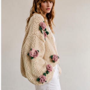 May include: Cream-colored, chunky knit cardigan adorned with pink rose appliqu&eacute;s and green leaves. The oversized sweater features a relaxed fit and is a cozy, stylish garment.