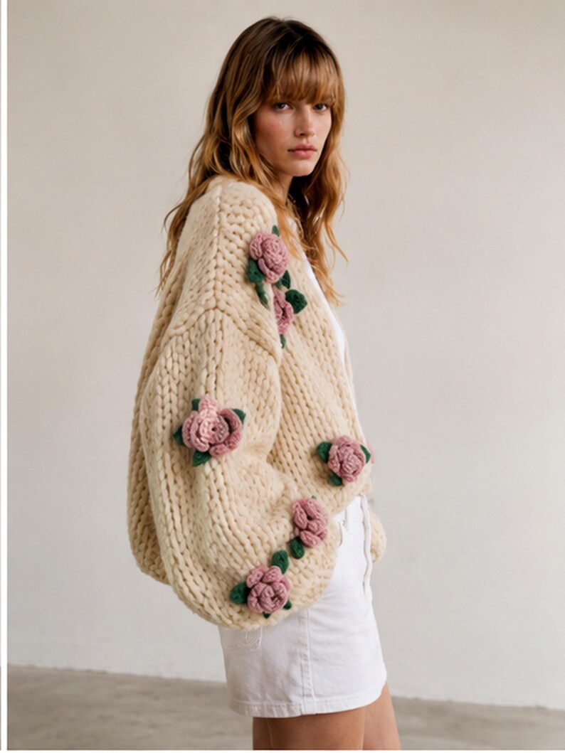 May include: Cream-colored, chunky knit cardigan adorned with pink rose appliqu&eacute;s and green leaves. The oversized sweater features a relaxed fit and is a cozy, stylish garment.