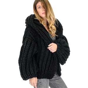 Op de afbeelding: Een grof gebreid zwart vest met een oversized capuchon, lange volumineuze mouwen en een dikke ribbelstructuur. Het kledingstuk is gemaakt van zwaar garen met een opvallend breipatroon, wat zorgt voor een robuuste en getextureerde uitstraling.