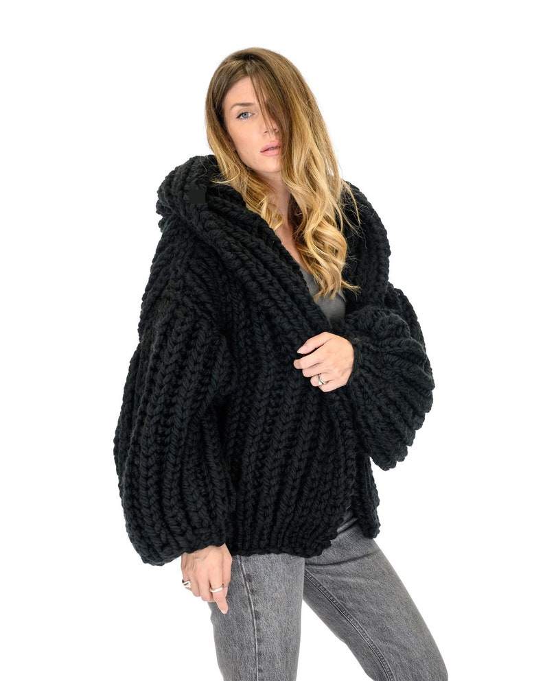 Op de afbeelding: Een grof gebreid zwart vest met een oversized capuchon, lange volumineuze mouwen en een dikke ribbelstructuur. Het kledingstuk is gemaakt van zwaar garen met een opvallend breipatroon, wat zorgt voor een robuuste en getextureerde uitstraling.
