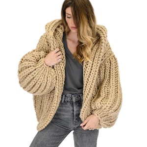 Op de afbeelding: Een grof gebreid, beige, oversized vest met een capuchon, lange mouwen en een dikke, geribbelde textuur. Het kledingstuk wordt gedragen over een donkergrijs T-shirt met V-hals en een antracietgrijze spijkerbroek, met zwaar garen.