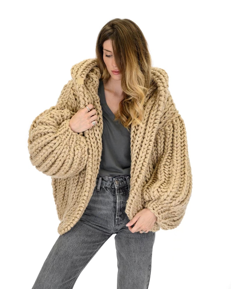 Op de afbeelding: Een grof gebreid, beige, oversized vest met een capuchon, lange mouwen en een dikke, geribbelde textuur. Het kledingstuk wordt gedragen over een donkergrijs T-shirt met V-hals en een antracietgrijze spijkerbroek, met zwaar garen.
