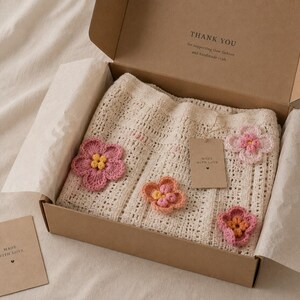 Pu&ograve; includere: Un capo all'uncinetto color crema con applicazioni floreali rosa e arancioni, presentato in una scatola di cartone marrone. La scatola include un piccolo tag che dice "Made with Love". Il coperchio della scatola ha il testo "Thank You".