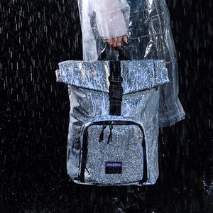 May include: A person wearing a clear raincoat and carrying a silver backpack in the rain. The backpack has a roll-top closure, a front pocket, and black straps. The background is dark, with visible raindrops.