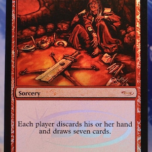 May include: A red Magic: The Gathering card titled "Wheel of Fortune." The card features an illustration of a robed figure and a sword. Text on the card reads "Each player discards his or her hand and draws seven cards."