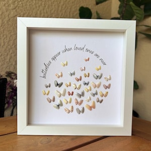 May include: A square white frame containing a white canvas with the black text "butterflies appear when loved ones are near" arranged in a semi-circle above a cluster of small, multi-colored three-dimensional butterfly embellishments.