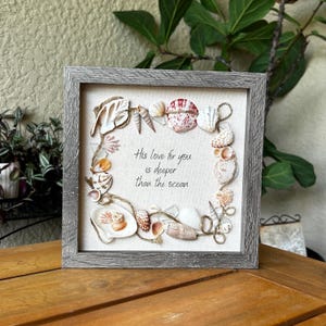 May include: A square wall art piece in a gray wood-grain frame featuring a cream-colored fabric background. A border of various seashells and twine surrounds the black printed text: His love for you is deeper than the ocean.