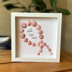 May include: A framed artwork featuring a white background with a design made of seashells. The seashells are arranged in a ribbon shape, with the words "faith hope cure" in the center. The frame is white.