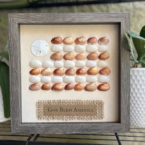 May include: Framed artwork featuring a flag design made of seashells. The flag includes a white sand dollar and rows of brown and white shells. The phrase "God Bless America" is displayed on a burlap banner. The frame is gray.