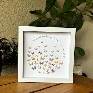 May include: A square white frame containing a white background with numerous small, multi-colored butterfly-shaped embellishments arranged in a cluster. The text "For He will order His angels to protect you wherever you go" and "Psalm 91:11" is printed in black.