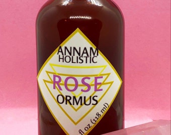 Könnte beinhalten: Eine braune Glasflasche mit schwarzem Deckel, beschriftet mit "ROSE ORMUS" von "ANNAM HOLISTIC". Das Etikett ist weiß und goldfarben mit einer rautenförmigen Form. Die Flasche enthält 118 ml. Ein rosa Kristall befindet sich im Vordergrund vor einem rosa Hintergrund.