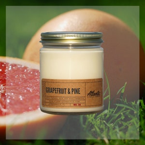 May include: A clear glass jar candle with a gold lid. The candle is labeled "GRAPEFRUIT & PINE" and "Atlantic Candle Company". The candle is sitting on a bed of green grass.