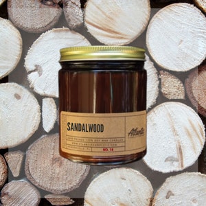 May include: A brown glass candle jar with a gold lid. The label reads "Sandalwood" and "Atlantic Candle Co.". The candle is sitting on a pile of wooden logs.