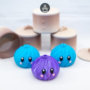 May include: Three small, round, swirled dumpling-shaped figures with large black eyes and pink cheeks, one purple and two blue, positioned in front of three beige cylindrical containers with lids, one of which is open and lying on its side.