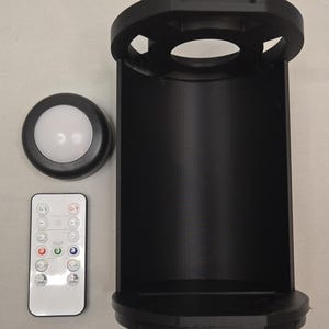 May include: A black cylindrical light fixture housing with an open front, a circular white LED puck light, and a rectangular white remote control featuring buttons labeled ON, OFF, color options, and timer settings for 15, 30, and 60 minutes.