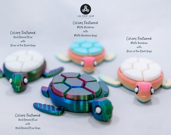 Puede incluir: Cuatro juguetes fidget con forma de tortuga de colores. Las tortugas presentan una variedad de colores, incluyendo arcoíris mate, rojo, verde y azul. El texto de la imagen dice: "Colores: Rojo/Verde/Azul con teclas que brillan en la oscuridad."