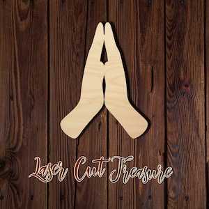 May include: Wooden cutout of two hands clasped together in a prayer position. The cutout is made of light brown wood and is set against a dark brown wood background.