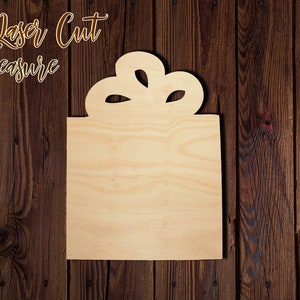 May include: A wooden cutout of a gift box with a bow on top. The cutout is unfinished and ready to be painted or stained.