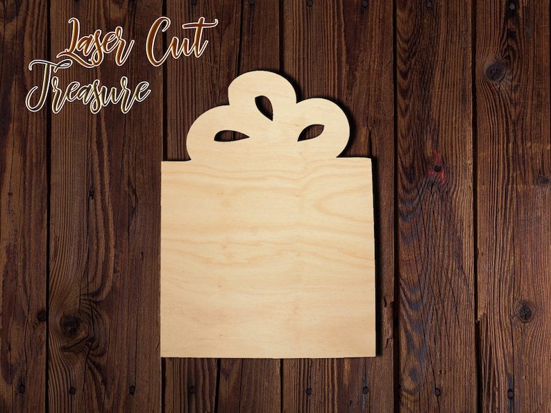 May include: A wooden cutout of a gift box with a bow on top. The cutout is unfinished and ready to be painted or stained.