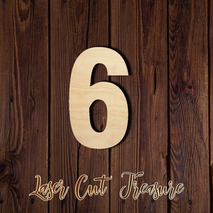 May include: A wooden number six cutout against a dark brown wood background. The number is light brown and has a smooth surface. The text "Laser Cut Treasure" is written in cursive below the number.