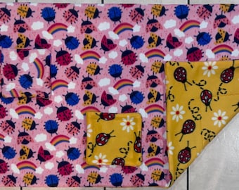 May include: A pink and yellow reversible blanket with a ladybug and rainbow pattern on one side and a yellow floral pattern on the other side. The blanket has two pockets on the pink side.