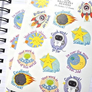 May include: A sheet of colorful space-themed stickers with phrases like "Brilliant," "Excellent," and "Stellar job!" The stickers feature rockets, planets, stars, and smiling characters. A blue and white pen and a spiral notebook are in the background.