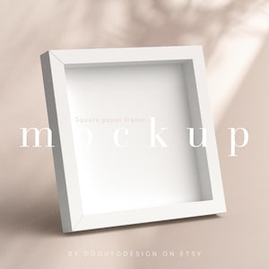 May include: White square picture frame mockup on a light beige background. The frame is sitting on a surface with shadows from an overhead light source. The text "Square paper frame mockup" is visible in the frame.