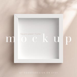 Puede incluir: Maqueta de marco de papel cuadrado blanco sobre un fondo beige claro. El marco está sentado en una superficie con sombras de una ventana o fuente de luz. El texto "Square paper frame mockup" está escrito en letras blancas en el marco.