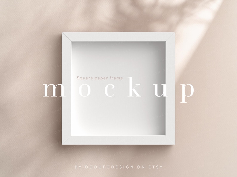 May include: White square paper frame mockup on a light beige background. The frame is sitting on a surface with shadows from a window or light source. The text "Square paper frame mockup" is written in white letters on the frame.