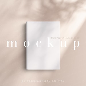 Puede incluir: Maqueta de libro-diario de tapa dura blanca sobre fondo beige con la palabra "mockup" en fuente clara. El texto "Hard Cover Journal-Book" está encima del diario y "BY DODURODESIGN ON ETSY" debajo.