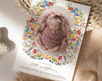 May include: A white card with a watercolor floral wreath surrounding a photo of a baby girl. The text "introducing our little wildflower" and "CHARLOTTE ELAINE MARTELL" is printed below the photo. The card is on a woven wicker surface.